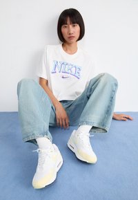 Vit Nike-t-shirt med lila och blå logotyp, ljusblå baggy jeans och vita sneakers med gula detaljer, sittande på en blå yta.