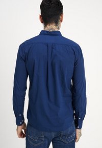 Námořnická modrá košile s dlouhým rukávem, bavlněný materiál, manžety na knoflíky, design s průhybem na zádech. Hladká textura bez viditelných vzorů nebo akcentů.