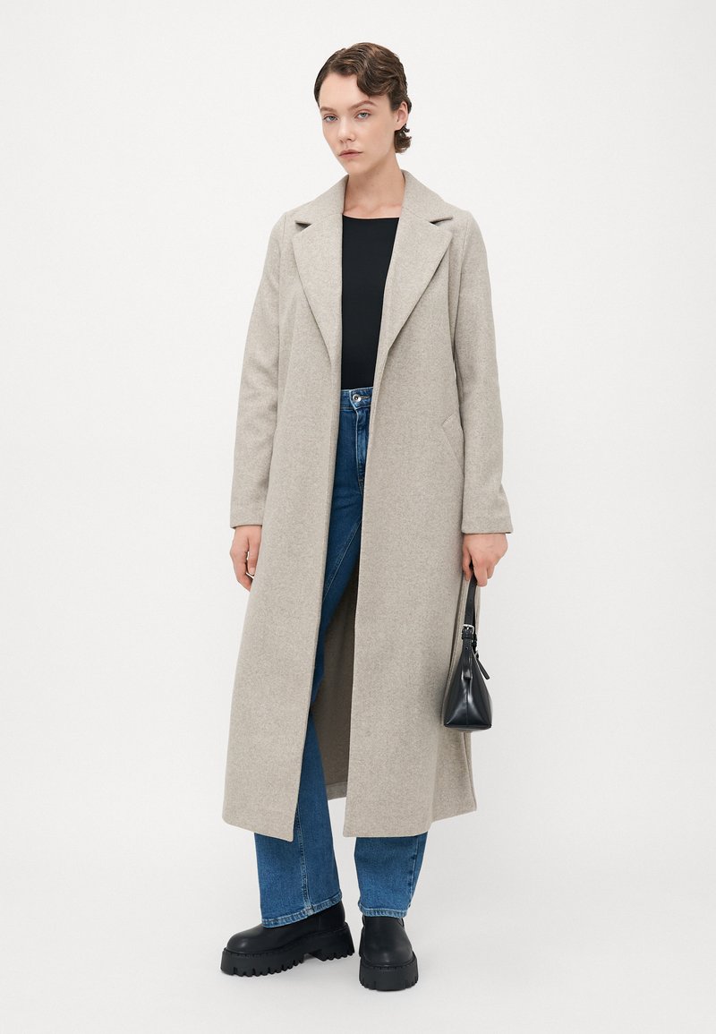 Femme portant un long manteau beige sur un haut noir et un jean bleu, tenant un petit sac à main noir, se tenant devant un fond blanc uni.