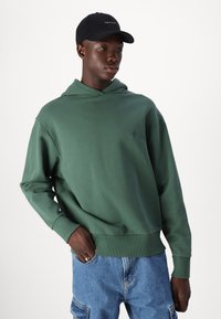 Grønn bomullshoodie med avslappet passform, ribbestrikkede mansjetter og hette med snøring. Bært med blå denimshorts, som viser et sporty design.