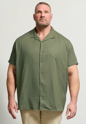 Muskelbepackter Mann mit kurzen Haaren und Bart, der ein lockeres olivgrünes kurzärmeliges Hemd mit Knöpfen und beige Hosen vor schlichtem Hintergrund trägt.