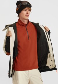 Fleece quarter-zip top in roestbruin, onder een zwart en crèmekleurige jas. Het model draagt een zwarte beanie en skibril. Textuurdetails van de stof zichtbaar.