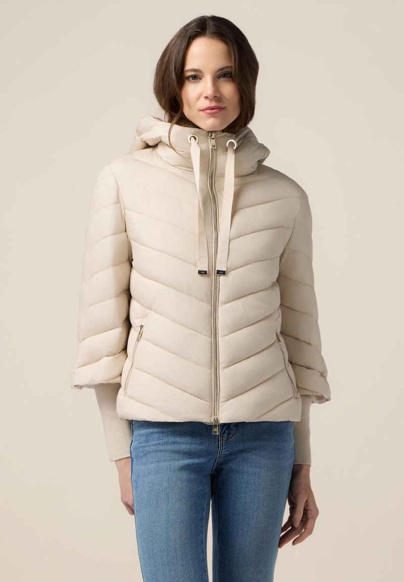 Beige pufferjakke med chevron-syninger, en hætte med snoretræk og lynlåslommer i siden. Har et cropped design og en blød tekstur.