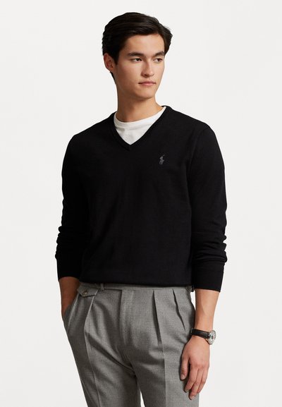 Schwarzer V-Ausschnitt-Pullover aus weichem Material, mit langen Ärmeln und einem kleinen Logodetail auf der Brust, kombiniert mit grauen, gefalteten Hosen.