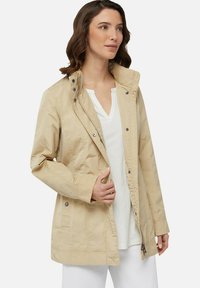 Giacca impermeabile beige chiaro con colletto alto, bottoni a pressione e chiusura con zip. Presenta tasche laterali e una vestibilità comoda.