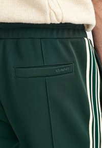 Zielone spodnie sportowe wykonane z gładkiego materiału, z tylną kieszenią z wytłoczonym logo "GANT" oraz białymi paskami po bokach nogawki.