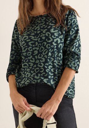 Mujer con una blusa de manga larga de estampado de leopardo en verde oscuro y negro con las mangas remangadas, sosteniendo un bolso beige claro frente a unos pantalones negros.