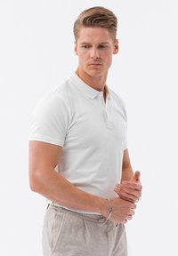 Bílá polo košile s krátkým rukávem a límečkem, s knoflíkovou légou. Model má na sobě svetrové kalhoty světle béžové barvy a stříbrný náramek.