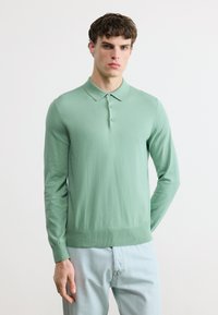 Jeune homme portant un polo manches longues vert menthe et un jean bleu clair, debout avec une main dans la poche sur un fond blanc uni.