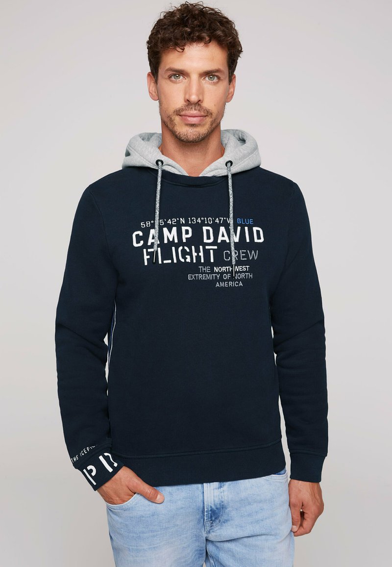 Sudadera azul marino con capucha gris, con el texto "CAMP DAVID FLIGHT CREW". Incluye cordones y puños acanalados. Tejido de algodón suave.