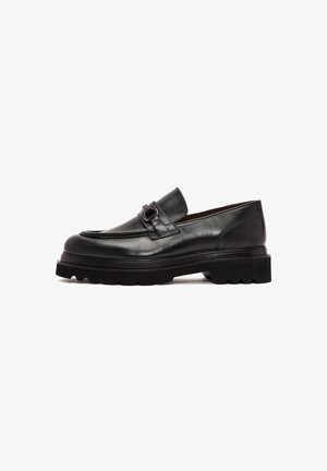 Mocasines de cuero negro con punta redondeada, una correa decorativa con herrajes metálicos y una suela negra gruesa con relieve para mayor agarre.