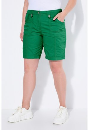 Person iført grønne knælange shorts med knapdetaljer og hvide sneakers, stående mod en ensfarvet hvid baggrund.