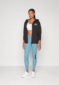 Čierna mikina na zips s logom, biely crop top a svetlomodré legíny s bočným logom. Biele športové topánky. Hladká textúra látky.