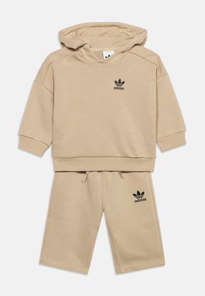 Béžový detský outfit Adidas so svetrom s kapucňou a zodpovedajúcimi šortkami, obe s čiernymi logami Adidas trefoil na ľavej strane.