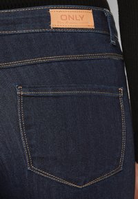 Mørkeblå denimjeans med en sydd baklomme og en lærmerkelapp, med kontrasterende gult søm langs sømmene.