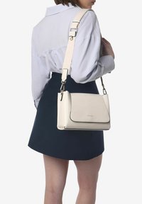 Bolso cruzado de piel blanca con cierre de solapa, correa ajustable y sutiles detalles de costura. Textura suave y forma rectangular.
