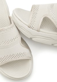 Beige sandals with knit fabric straps, a textured sole, and a contoured footbed. Open toe design with elastic accents for comfort.