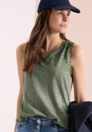 Femme portant un haut sans manches vert, un jean bleu en denim avec boutons, et une casquette bleu foncé, tenant une veste bleu marine d’une main.