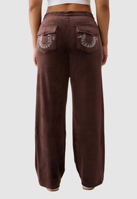 Osoba ubrana w brązowe spodnie z szerokimi nogawkami, z klapami na tylnej kieszeni ozdobionymi wzorami z kryształków, pokazana z tyłu.