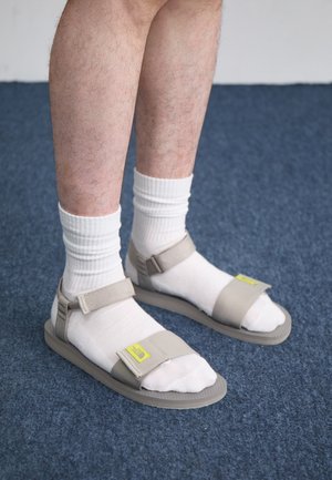 Des sandales beiges à brides réglables, portées sur des chaussettes blanches. Les semelles sont plates avec un motif texturé. Sur fond de moquette bleue.