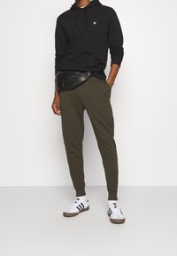 Felpa nera con logo giallo, pantaloni jogger verde oliva, borsa a tracolla nera e sneakers bianche con strisce nere. Abbigliamento casual sportivo.