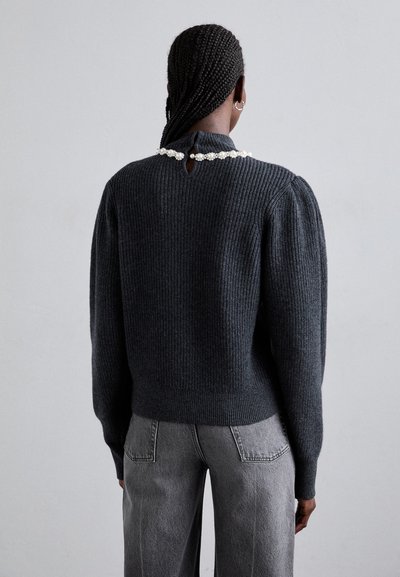 Femme aux cheveux tressés portant un pull côtelé foncé avec un détail en perles à l'encolure arrière et un jean gris, de dos contre un fond uni.
