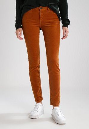 Person iført slanke rustfarvede manchesterbukser med hvide sneakers og en sort trøje, stående mod en ensfarvet baggrund.