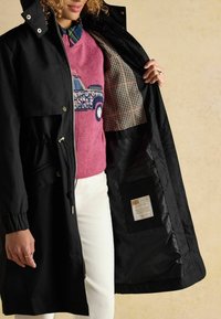 Čierna dažďová bunda so zipsami, sťahovacím pásom a kockovanou podšívkou. Interiér obsahuje vrecko s etikietami. Model má na sebe ružový sveter.