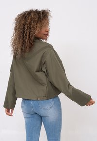 Olivgrüne, kurze Jacke mit glatter Textur, weitem Schnitt an den Ärmeln und Knopfdetails am Saum. Kombiniert mit hellblauen, eng anliegenden Jeans.