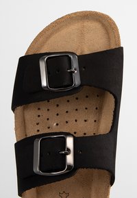 Sandale en cuir noir avec deux boucles ajustables, une semelle intérieure en liège texturé et des perforations pour la ventilation. Conçue pour le confort et le soutien.