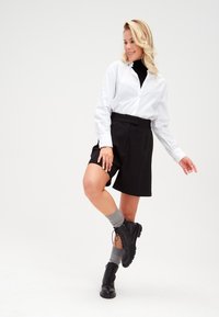 Camicia bianca con bottoni e pantaloni corti neri a pieghe, abbinati a calzini grigi e stivali neri stringati, su uno sfondo neutro.