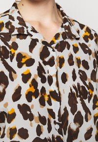 Leopardmönstrad skjorta i lätt tyg; har bruna, krämfärgade och gula fläckar med klassisk krage och vita knappar på framsidan.