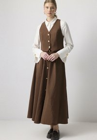 Vestido largo marrón con un top tipo chaleco ajustado y un diseño de botones en la parte delantera, superpuesto a una camisa blanca con cuello. Textura suave, diseño elegante, combinado con zapatos oscuros.