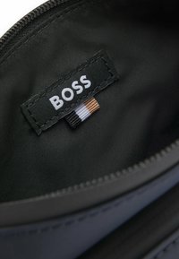 Interno di una borsa in tessuto nero caratterizzato da un evidente etichetta "BOSS", completato da un'etichetta testurizzata con accenti marroni, bianchi, neri e argentati.