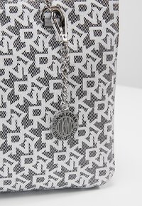 Gros plan d’un coin de sac à main texturé noir et blanc avec des lettres "DKNY" répétées et un pendentif chaîne argentée indiquant "Donna Karan - DKNY - 1989."