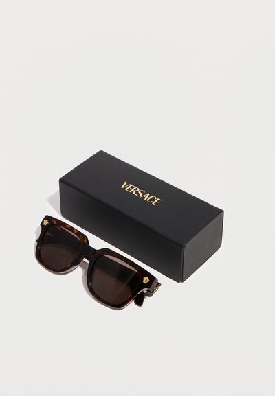 Σκούρα γυαλιά ηλίου με τετράγωνα σκελετά σε σχέδιο χελώνας και χρυσά λογότυπα Medusa δίπλα σε μαύρη ορθογώνια θήκη Versace σε λευκό φόντο.