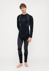 Zwarte sportieve basislaagset met een lange mouwen top met grijze accenten en aansluitende leggings met structuurpatronen. Glad, rekbaar materiaal.