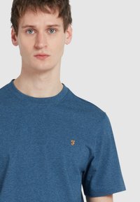 T-shirt in cotone blu con maniche corte, girocollo e un piccolo logo arancione sul lato sinistro del petto. Texture liscia, design casual.