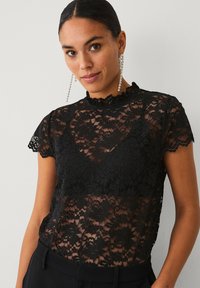 Sort bluse i blonde med korte ærmer, bølget udskæring og blomstermønstre. Båret med sorte bukser med høj talje. Glitrende øreringe.