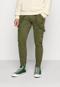 Pantalons cargo vert olive avec plusieurs poches latérales, coupe ajustée, poignets élastiques et une texture de tissu. Portés avec des baskets montantes vertes.