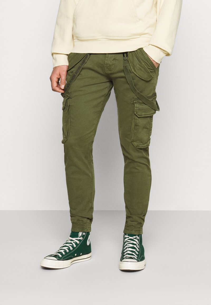 Pantalons cargo vert olive avec plusieurs poches latérales, coupe ajustée, poignets élastiques et une texture de tissu. Portés avec des baskets montantes vertes.