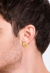 Orecchino in metallo dorato con un design ondulato e testurizzato. Forma circolare, caratterizzato da un piccolo cerchio con un'estremità aperta.