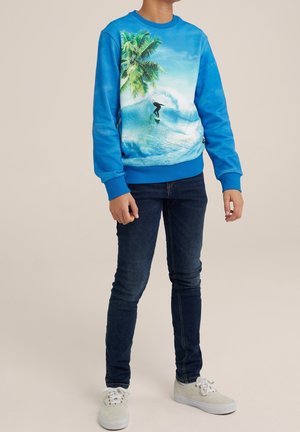 Dziecko w niebieskiej bluzie z tropikalną sceną surfingową, ciemnych dżinsach i białych trampkach, stojące na jednolitym tle.