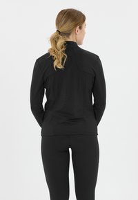 Sort langærmet atletisk jakke med høj krave, struktureret stof og sømme, der løber ned ad ryggen. Bæres med sorte leggings.