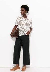 Haut en dentelle florale blanche, pantalon noir à jambes larges et baskets brunes avec des lacets exposés. Sac à main à motifs léopard marron et accessoires dorés.
