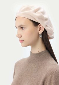 En mjuk rosa stickad basker ovanpå en modells huvud, i kombination med silverfärgade droppformade örhängen. Modellen bär en ljusbrun polotröja.