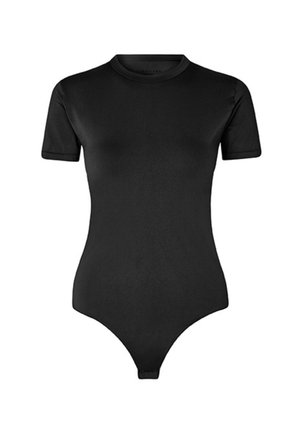 Kvinde med skulderlangt brunt hår iført en tætsiddende sort bodysuit med korte ærmer, stående mod en ensfarvet lys baggrund.