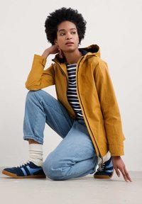 Geel waterdicht jack met een zwarte capuchon, gedragen over een gestreept shirt. Lichtblauwe relaxed-fit jeans en blauwe sneakers met gele accenten.