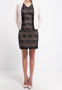 Abito in pizzo nero con ritagli geometrici sovrapposto a una sottoveste nera solida, abbinato a un blazer corto rosa chiaro e tacchi nudi.