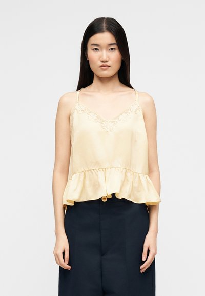 Femme aux longs cheveux noirs portant un caraco en soie jaune pâle avec des bordures en dentelle et un ourlet à volants, associé à un pantalon noir taille haute.